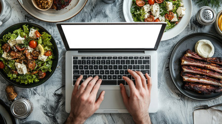 A white guy is typing on a laptop with a white screen, top view, close-up. Laptop mockup. There are plates of Greek cuisine on the table - lamb ribs and Greek salad. The concept of relaxation and enjoymentの素材