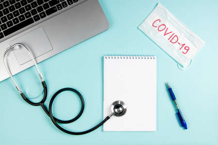 Flatlay doctor workplace with laptop, mask and notepad mock up. Text on medical mask "Covid-19"の写真素材
