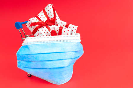 Shopping trolley with gifts in a protective face mask on a red background with copy space. The concept of buying gifts for the holiday during the virus.の写真素材