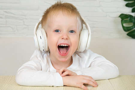 Little european boy smiles in a white t-shirt with big white headphones, listens to music and looks at the camera. The concept of music, home leisure.の写真素材