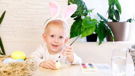 A little boy in rabbit ears enthusiastically paints Easter eggs with paint. Religious holidays concept, do it yourself, family leisure, easter egg huntの写真素材