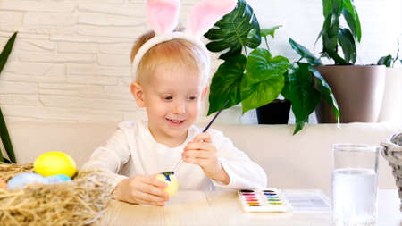 A little boy in rabbit ears enthusiastically paints Easter eggs with paint. Religious holidays concept, do it yourself, family leisure, easter egg huntの写真素材