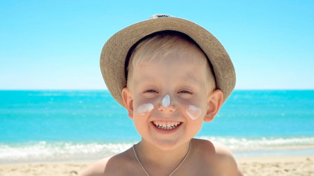 Positive emotional happy kid, smiling and laughing in a trendy hat, sunscreen on his face against the backdrop of sea waves. Rest, swim on the paradise ocean beach. Vacation and holidays with familyの写真素材