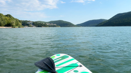 Paddleboard resting on tranquil water, with green and white design, surrounded by hills and blue sky, capturing the essence of outdoor leisure and adventureの写真素材
