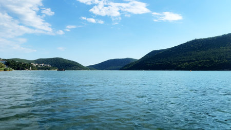 Serene landscape featuring calm blue water reflecting the sky, surrounded by lush green hills under a bright sunny day, creating a peaceful natural environmentの写真素材