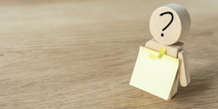 Wooden figure with a question mark on its head holds a blank yellow sticky note, conceptualizing asking, doubt, uncertainty, and seeking information or answersの素材
