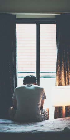 Man sitting alone on bed in a dark bedroom looking out window, representing feelings of loneliness, sadness, isolation, depression, anxiety, and contemplation during self-quarantineの素材