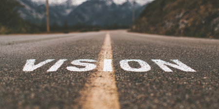 Vision word painted in white on a long grey asphalt road with a yellow dividing line, leading towards distant mountains, symbolizing future goals, journey, and strategic directionの素材