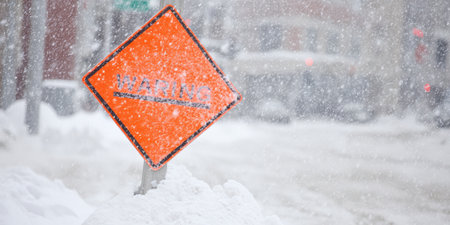 Orange warning sign standing in deep snow, with heavy snowfall impacting visibility and difficult driving conditions on a city street during a blizzardの素材