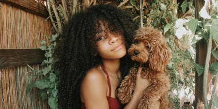 Young woman with beautiful long curly hair holding a cute brown poodle puppy outdoors, showing a genuine bond of friendship and companionship in a natural settingの素材
