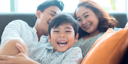 Close-up of an asian family of three embracing on a couch, parents smiling as their laughing young son looks at the camera, cozy living room bonding and joyの素材
