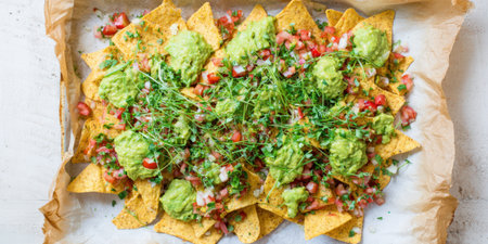 Nachos layered on baking paper topped with chunky guacamole, pico de gallo and fresh green sprouts for a colorful, crunchy party-ready appetizer or snackの素材