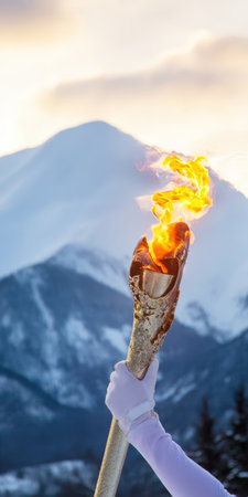 Athlete's hand wearing a white glove holding a blazing olympic torch, with a majestic snowy mountain range and a soft sky visible in the background, symbolizing sport, spirit, and winter gamesの素材