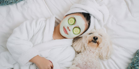 Woman wearing a face mask and cucumber slices on her eyes, relaxing comfortably on a bed next to her small white dog, enjoying a peaceful self-care beauty routine dayの素材