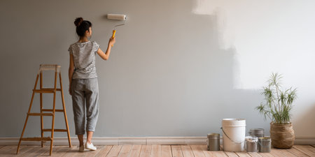 Young owner renovating and improving apartment, painting a gray wall with white color using a roller, standing near a wooden ladder and paint cans on the floorの素材