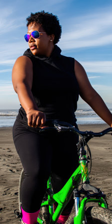 Black woman with short curly hair and purple sunglasses beside a green bicycle on sandy beach, gazing at calm blue ocean under clear sunny sky, enjoying active summer escapeの素材
