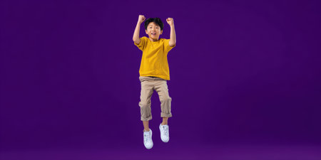 Happy asian boy jumping high in mid-air, raising fists, exuding joy and energy against a plain purple background, symbolizing achievement, freedom, and triumphの素材