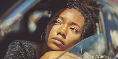 African american woman resting her head on the car window, looking thoughtfully into the distance, with sunlight casting shadows on her face while travelingの素材