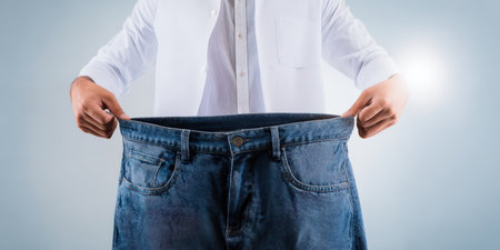 Man holding up his oversized jeans, demonstrating successful weight loss and health achievements after a diet and fitness program, symbolizing progress and a healthier lifestyleの素材