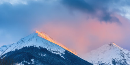 Snowcapped mountain peaks stand under a vibrant alpenglow sky with soft clouds, showcasing rugged slopes covered in snow and dark evergreen forests during a winter sunsetの素材