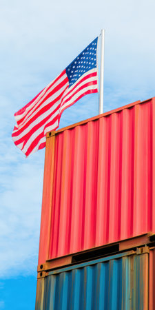 United states flag waving on a pole above two stacked cargo containers representing international trade, global logistics, import, and export against a blue skyの素材