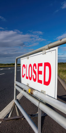 Metal barrier with a white closed sign in bold red letters blocking a rural highway, indicating no access or a temporary restriction under a clear blue skyの素材