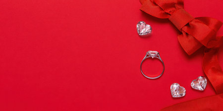 Engagement ring with a gemstone, shiny decorative hearts, and a red ribbon bow placed on a vibrant red background, symbolizing love, romance, and special occasionsの素材