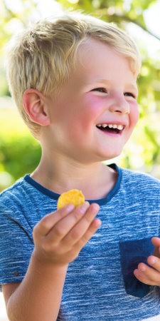 Blond boy smiling and laughing outdoors, holding a potato chip and enjoying a sunny, carefree summer snack moment, natural expression and joyful childhood leisureの素材