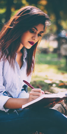 Young pensive woman sitting outdoors on green grass, focused on writing in her notebook with a pen, capturing ideas, journaling, or studying in a peaceful natural environmentの素材