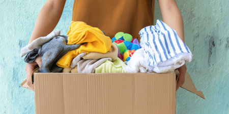 Person holding and offering a cardboard box filled with donated clothing and colorful toys, symbolizing charity, community support, reuse, and volunteer-driven generosity and aidの素材