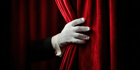 Magician's hand in a white glove pulling back a luxurious red velvet stage curtain, revealing darkness and anticipating a grand performance, show, or presentationの素材