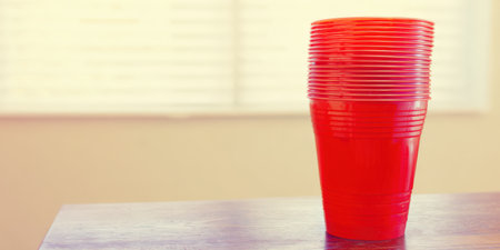 Stack of vibrant red disposable plastic cups standing on a dark wooden table, symbolizing party events, celebrations, drinking games, and social gatheringsの素材