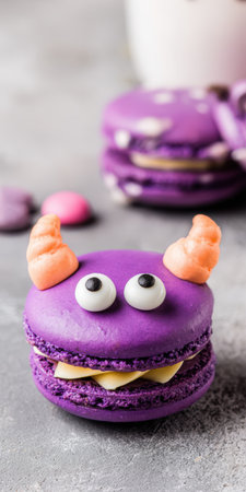 Purple macaron decorated with candy eyeballs and orange meringue horns as a cute monster treat, light filling visible on textured gray background ready for halloween partyの素材