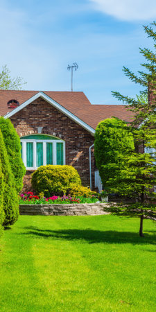 Suburban brick house featuring a well-maintained green lawn, vibrant garden beds with colorful flowers, and neatly trimmed hedges under a clear blue skyの素材