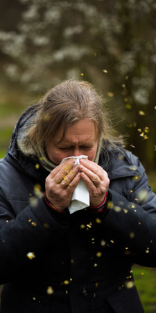 Woman feeling seasonal hay fever symptoms, sneezing into a tissue, surrounded by falling pollen in a natural outdoor environment, struggling with springtime allergiesの素材