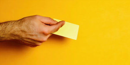 Hand holding a blank yellow payment card against a matching yellow background, providing clean copy space for branding, text or product and financial marketing conceptsの素材