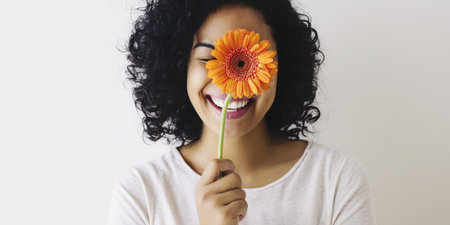Happy young woman with dark curly hair smiling broadly while holding a bright orange gerbera over one eye, playful studio portrait conveying joy, carefree summer energyの素材