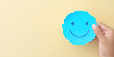 Hand holding a torn piece of blue paper with a drawn smiling face, symbolizing happiness, positivity, and a good mood on a light yellow background with copy spaceの素材