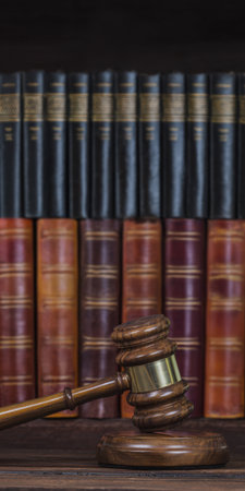 Judge gavel with a gold band resting on a wooden desk in front of a shelf of old law books, evoking justice, legal authority, courtroom decisions and vintage jurisprudenceの素材
