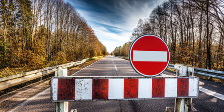 Red and white striped barrier with a red no entry sign blocking a rural road lined with trees under a blue sky, symbolizing restriction, prohibition, and an obstacleの素材