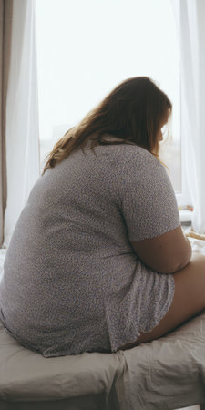 Overweight woman sitting on bed by a window, looking down with a pensive, lonely expression, conveying body image struggles, anxiety, low self-esteem and emotional distressの素材