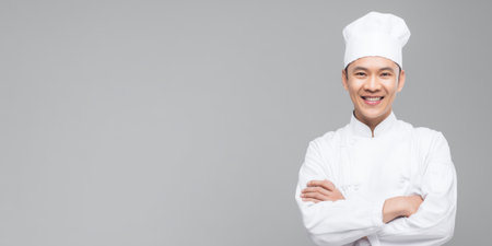 Asian male chef in white uniform and toque, standing with arms crossed and smiling confidently against a clean gray studio background, professional culinary portraitの素材