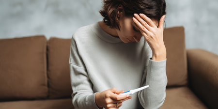 Woman sits alone on sofa looking down, holding a pregnancy test with a negative result, visibly upset and disheartened while struggling with infertility and trying to conceiveの素材