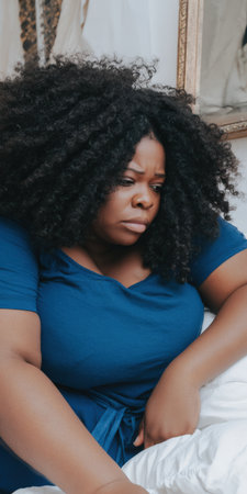 Plus size african american woman sitting indoors, feeling sad and anxious, struggling with emotional pain, body image issues, and mental health challengesの素材