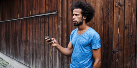Young man with an afro hairstyle holding a mobile phone in his hand, standing in front of a wooden wall and thinking while looking away, representing innovation, connection, and social mediaの素材