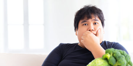 Overweight asian man feeling unhappy with a healthy diet, sitting on a sofa in a bright room and reluctantly looking at fresh broccoli, symbolizing struggles with weight loss and healthy eatingの素材