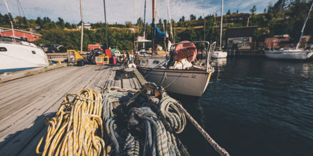 Mooring ropes coiled on a weathered wooden dock with sailboats bobbing in a tranquil harbor, sunlit water and blue sky evoking nautical leisure and coastal summer calmの素材