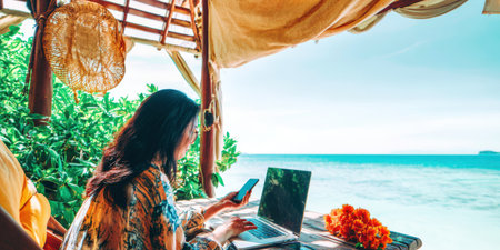 Woman focusing on her remote work and digital nomad lifestyle, typing on a laptop and using a smartphone from a shaded beach gazebo overlooking the clear blue oceanの素材