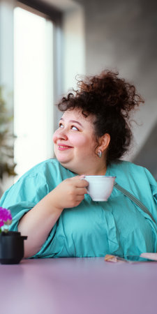 Plus size woman with curly hair smiling as she relaxes at a pink cafe table, enjoying a hot drink, embodying body positivity, contentment and peaceful morning leisureの素材