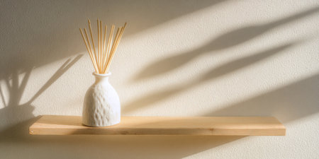 Reed diffuser with rattan sticks on a wooden shelf casting soft shadows on a textured off-white wall as warm sunlight fills a minimalist, calming interior space promoting wellnessの素材